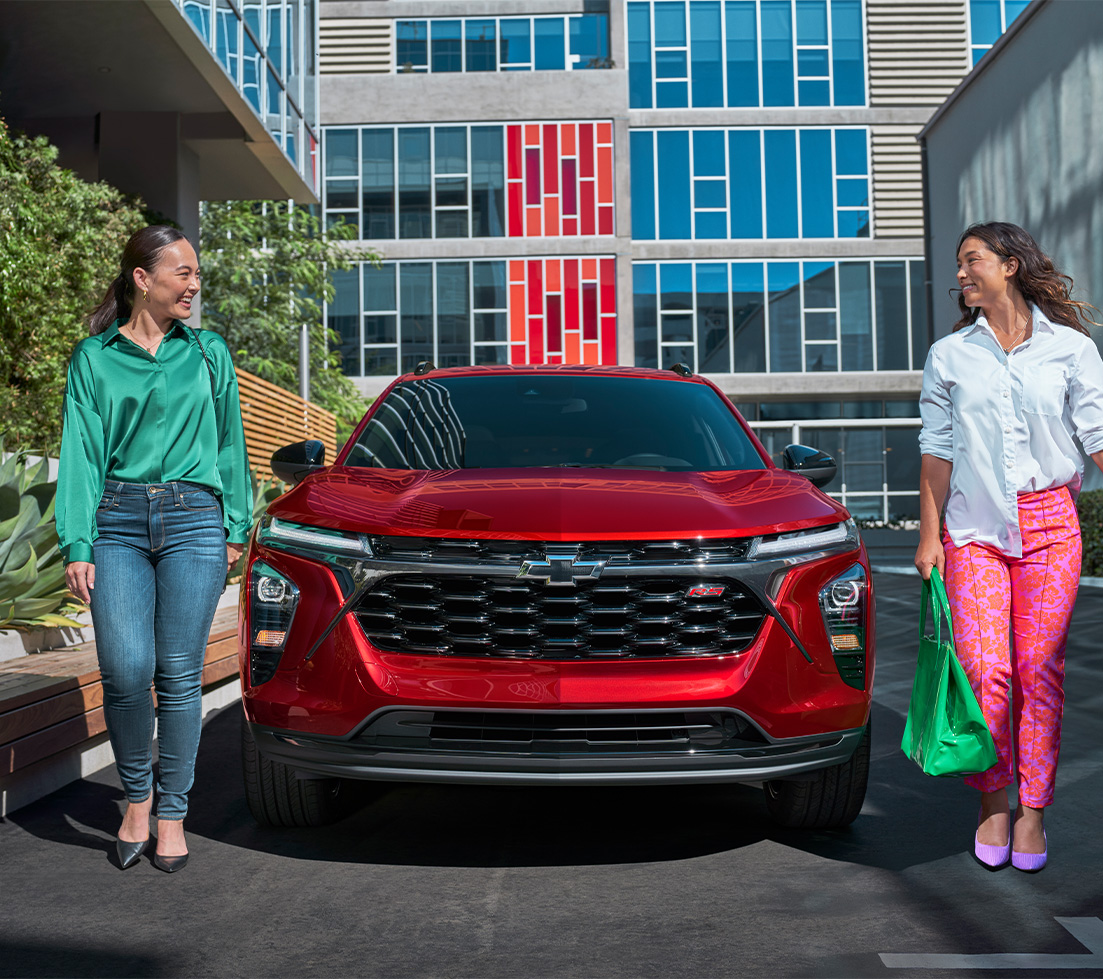 Two women are walking in front of a red Chevrolet Trax